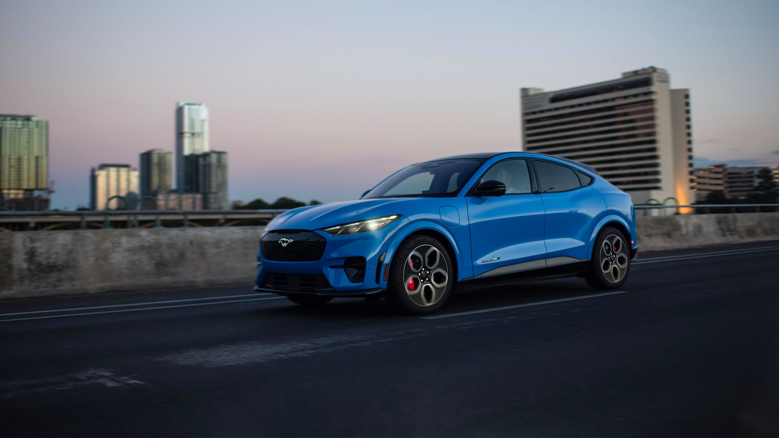 A blue 2024 Ford Mustang Mach-e drives along an urban highway 