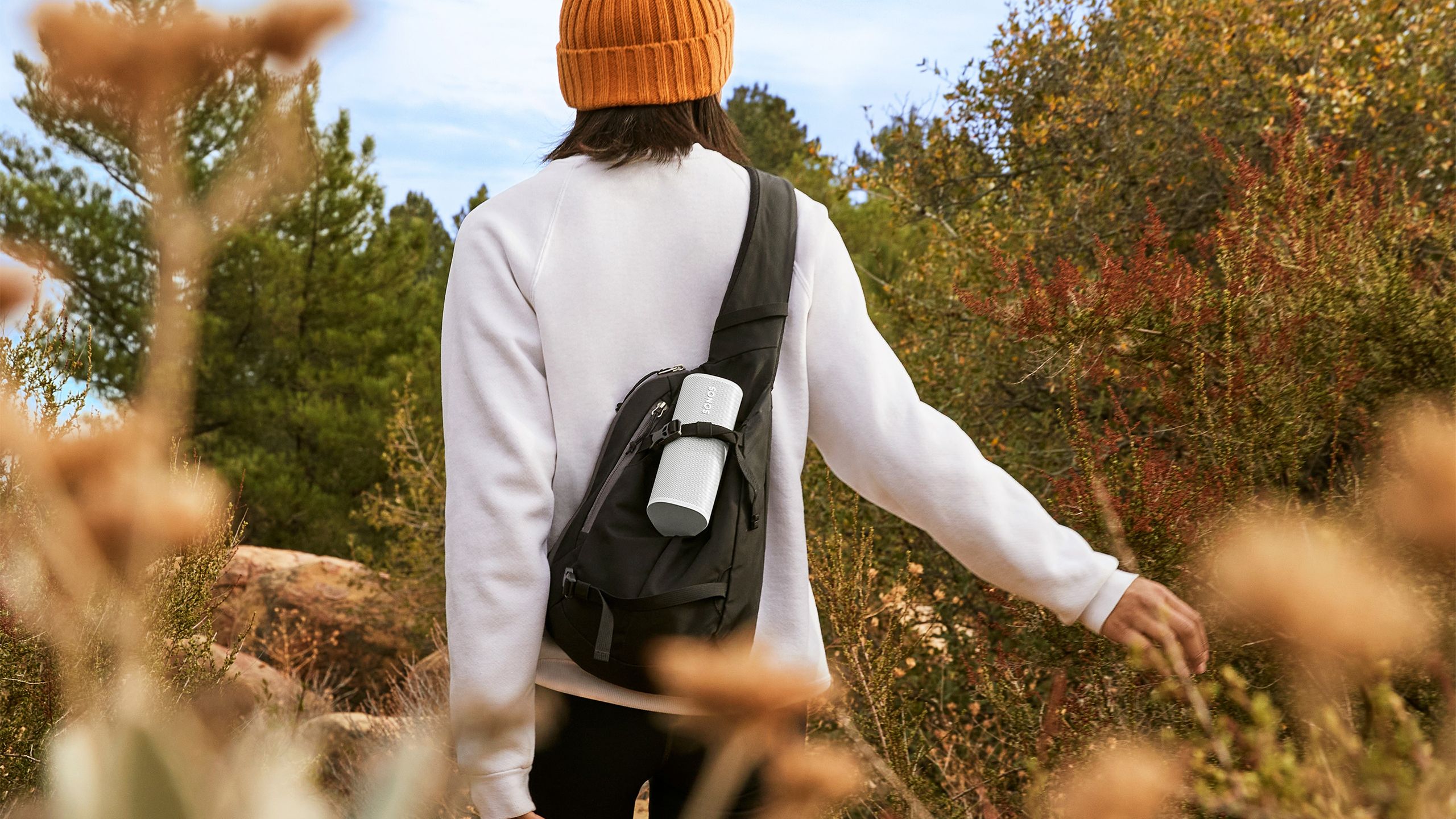 A person walks in a forest with the Sonos Roam 2 strapped to their backpack. 