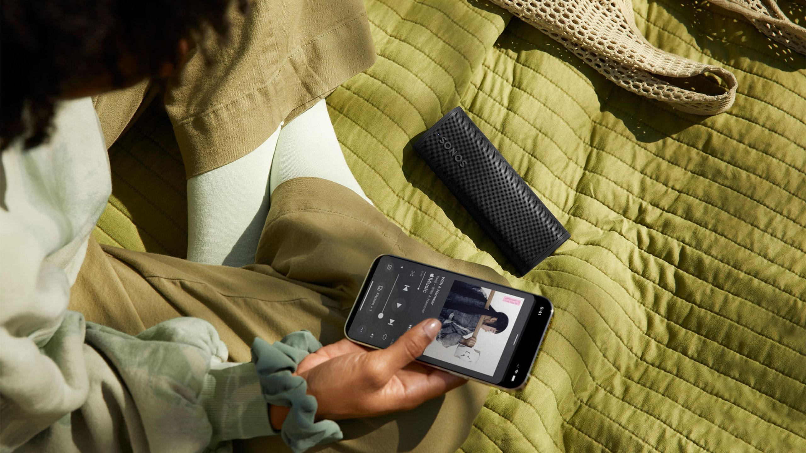 A person sits on a green blanket holding their phone next to the black Roam 2 speaker.