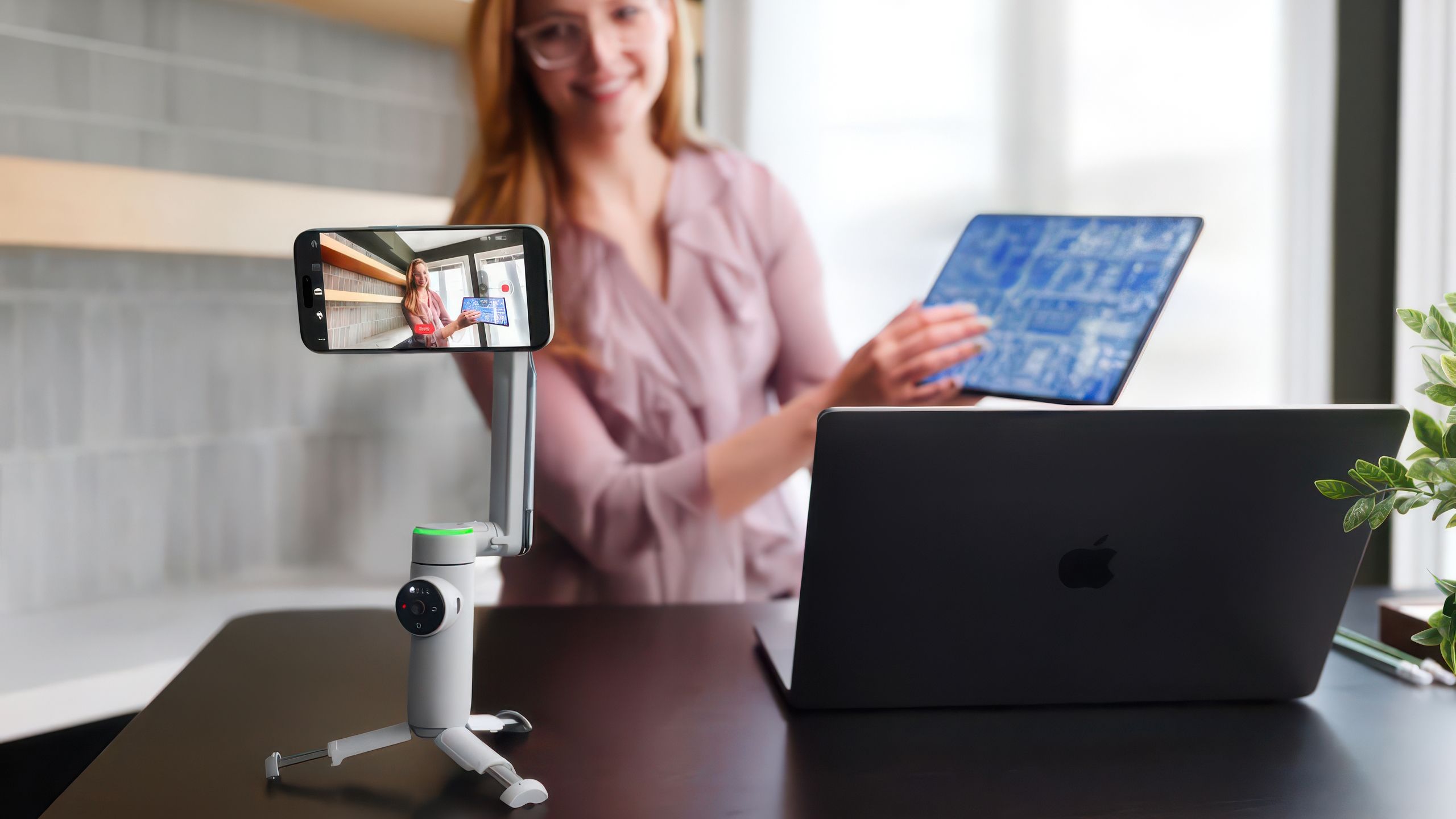 A woman stands at a desk with a computer and the Insta360 Flow Pro gimbal recording her.