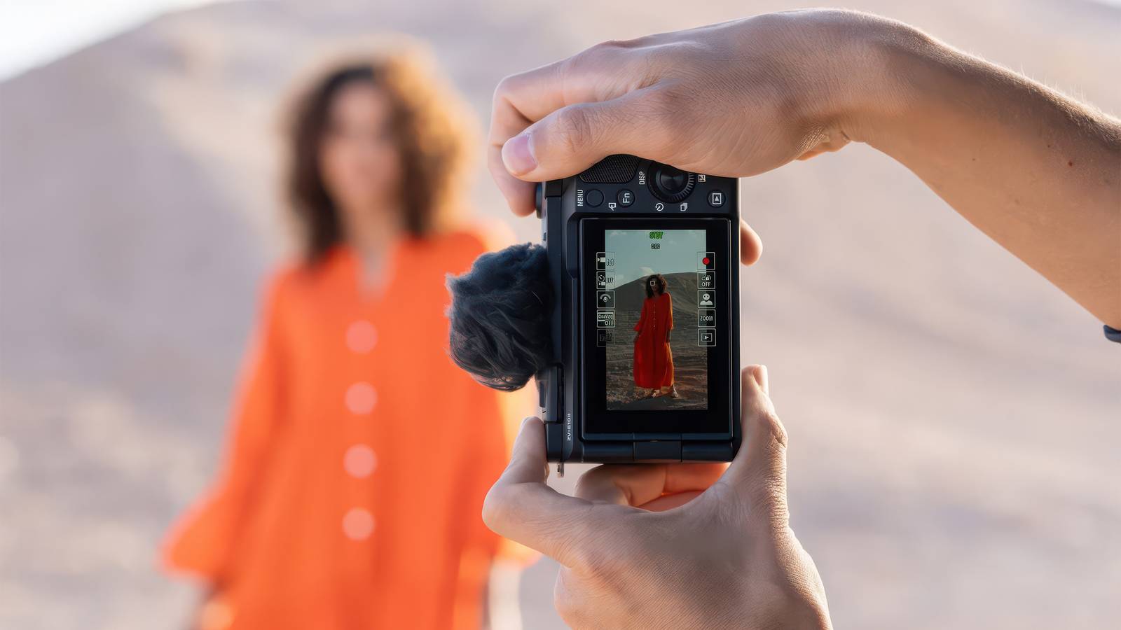 A pair of hands hold the Sony ZV-E10 II camera in front of a person with an orange dress.