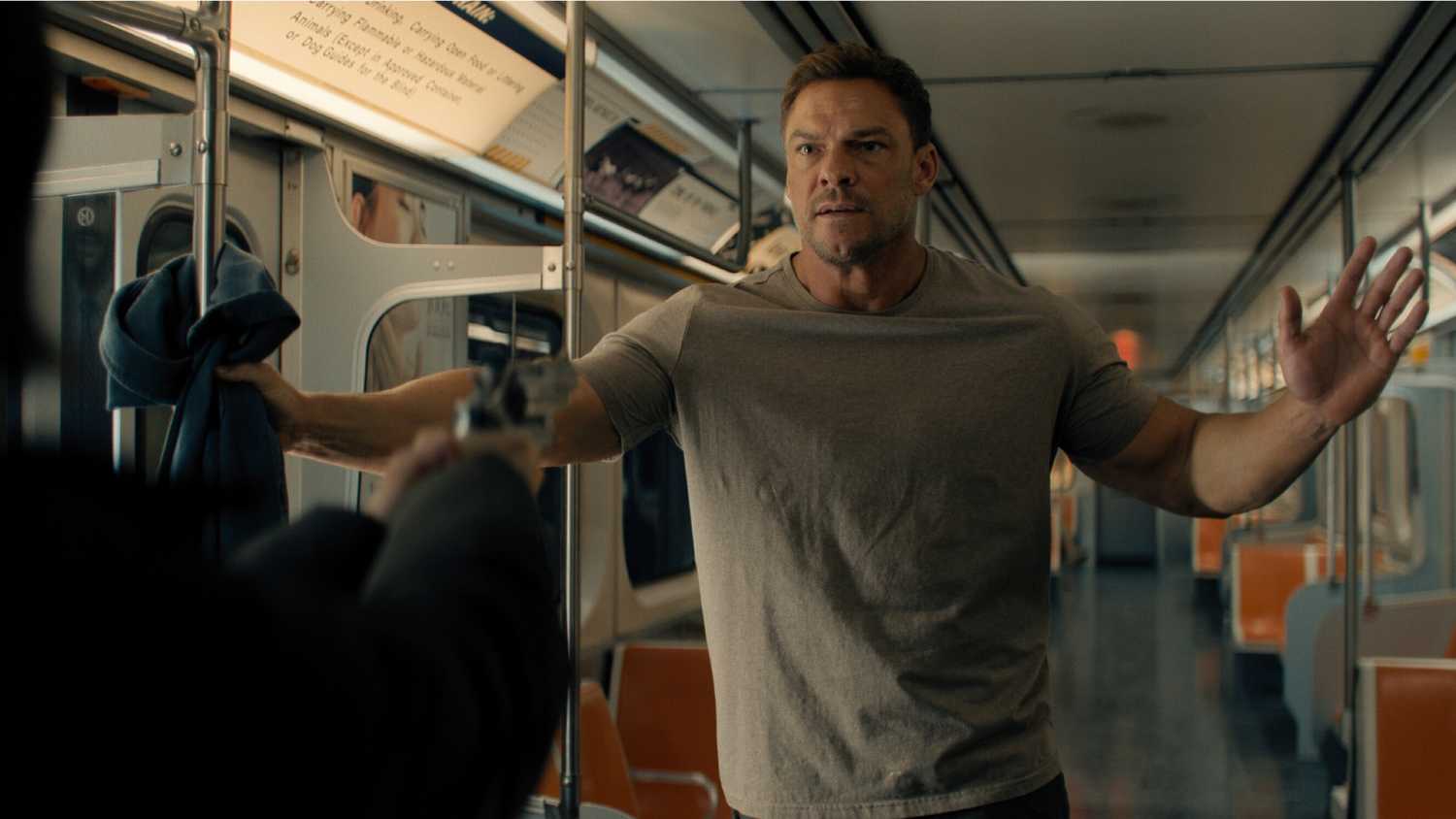 Alan Ritchson as Jack Reacher wearing a light brown shirt, with his hands in the air as someone points a gun at him.