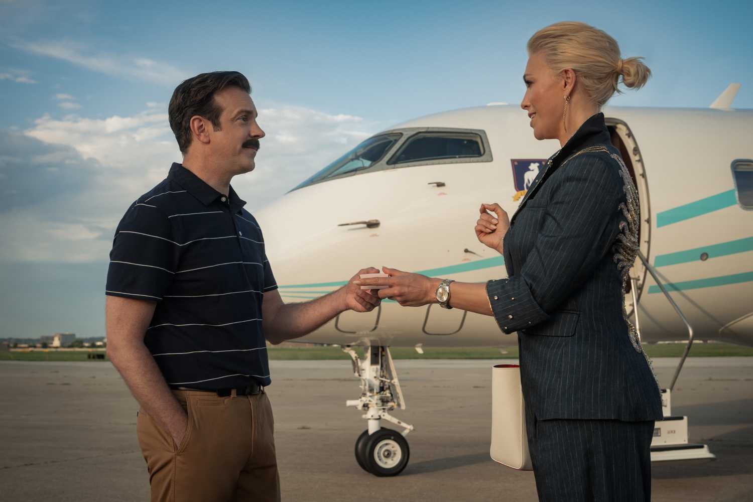Jason Sudeikis taking something from Hannah Waddingham with a plane in the background in Ted Lasso season 4.