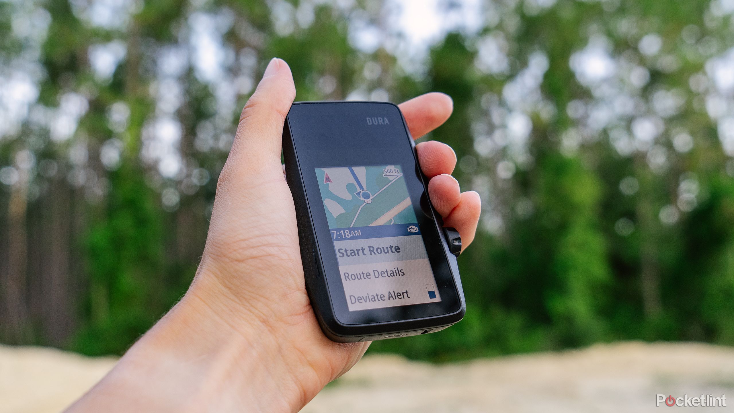 A hand holds the Coros Dura bike computer in front of a blurred out forest. 