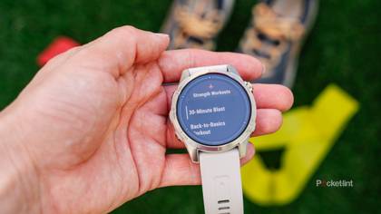 A hand holds the Garmin epix Pro Gen 2 Sapphire Edition above shoes and a yellow resistance band.