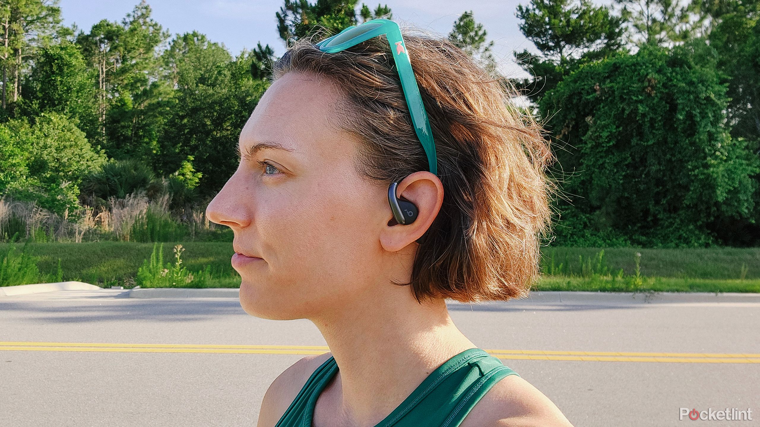 A woman wearing the Soundcore AeroFit headphones in front of a road with a forest background.