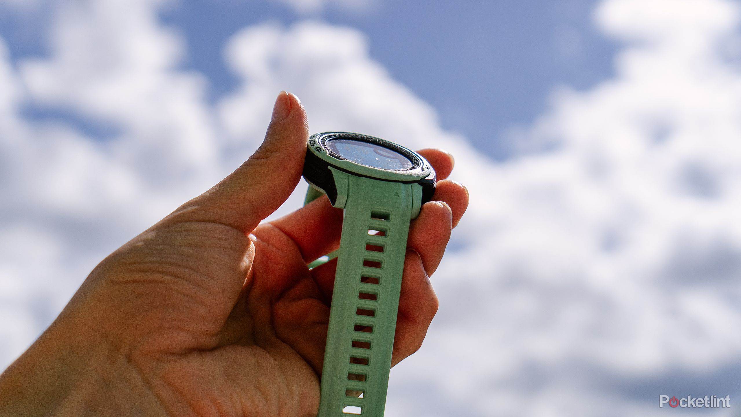 A hand holds the Garmin Instinct 2S Solar in front of blue sky with fluffy white clouds.