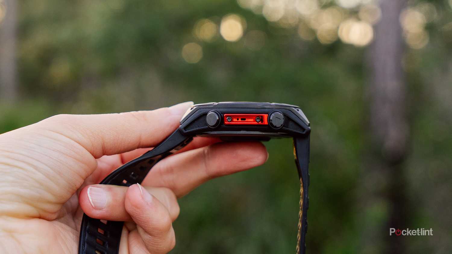 A watch holds the Garmin Fenix 8 with the speaker showing in front of blurred woods. 