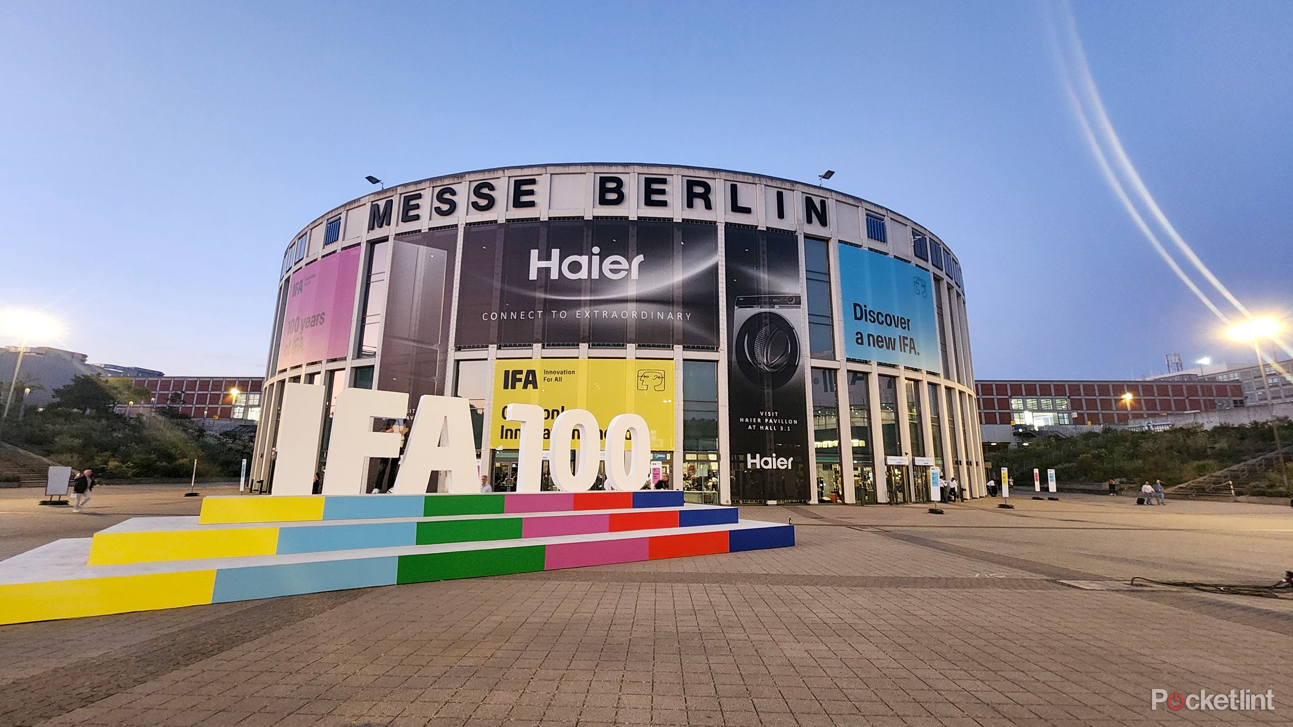Large letters that spell IFA 100 sit on colorful steps in front of a round building with advertising banners hanging on it. 