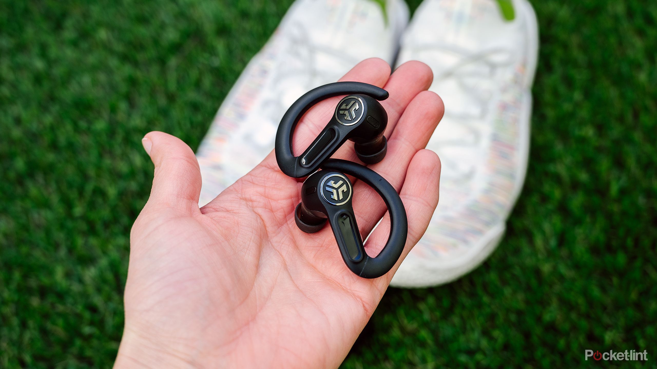 A hand holds the JLab Epic Sport ANC 3 Earbuds above a pair of shoes on green turf. 