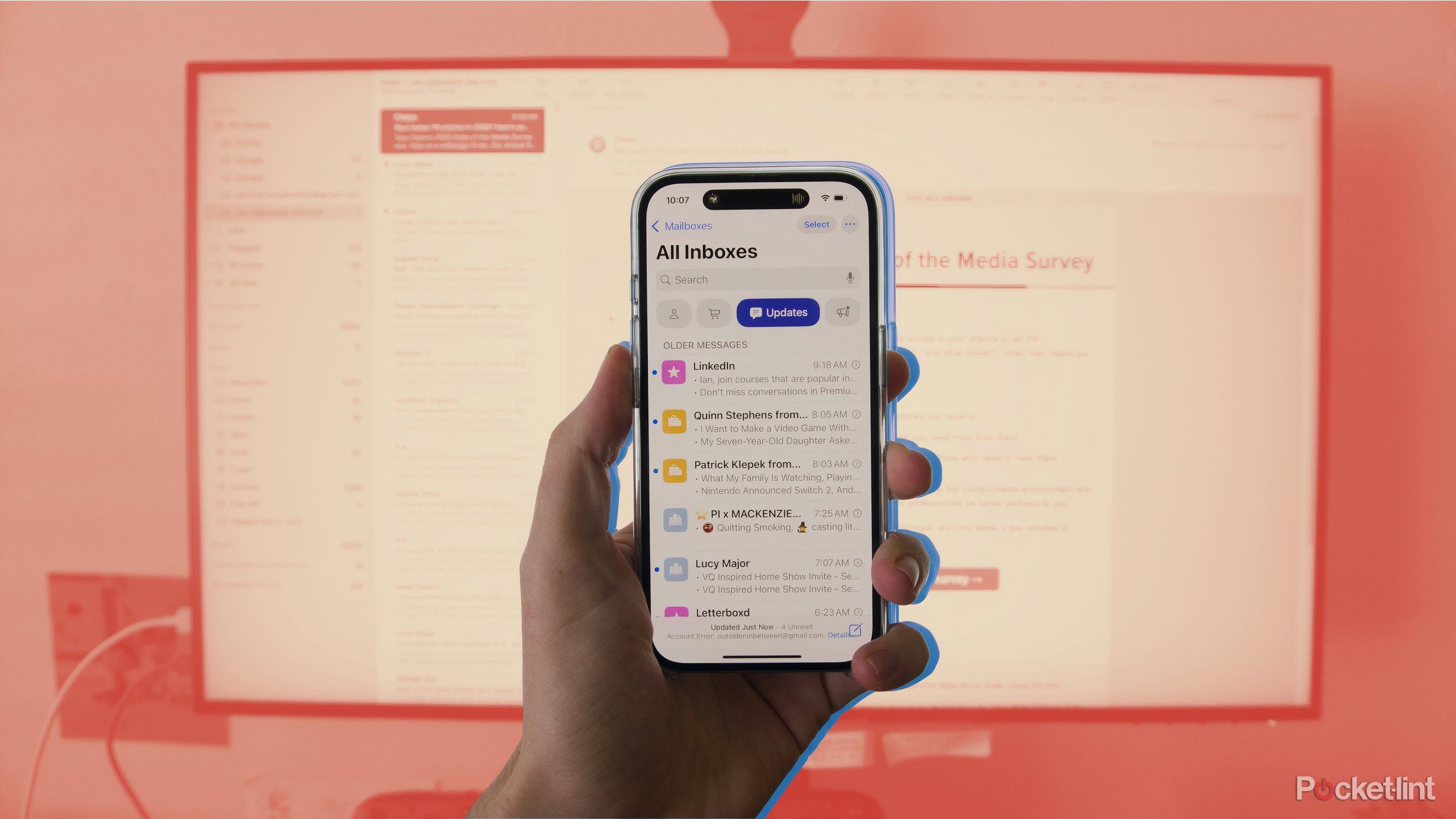 A hand holding an iPhone displaying the AI-powered Mail app in front of a screen showing macOS.