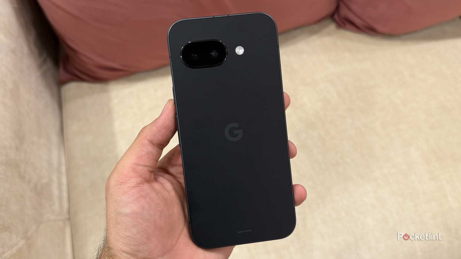 Google Pixel 9a in hand in front of a white sofa