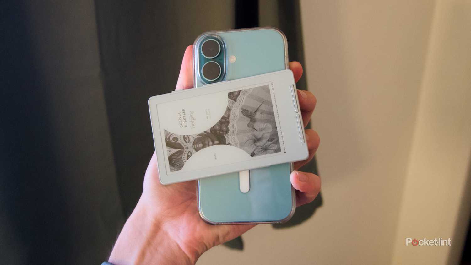 A hand holding an iPhone 16 with an Xteink X4 e-reader magnetically attached horizontally on its back.