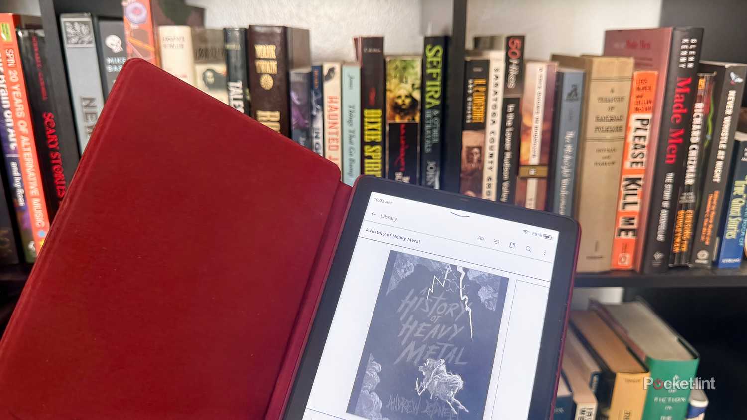 Kindle e-reader held in front of physical copies of books on shelf.